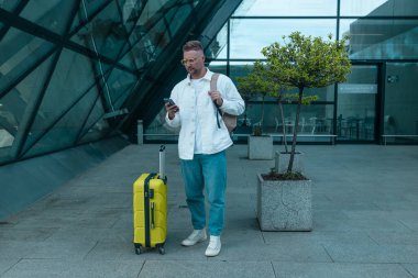 Akıllı telefon ve dizüstü bilgisayar kullanarak şık giyinen beyaz erkek yolcu, havaalanındaki dizüstü bilgisayarında eğitim toplantısı yapıyor. Uluslararası havaalanı transit havaalanı..