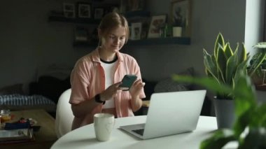 Genç kız öğrenci ev ödevini cep telefonu laptopuyla evde ağır çekimde hazırlıyor. Beyaz kadın akıllı telefondan proje hakkında bilgi arıyor. Serbest ev eğitimi 