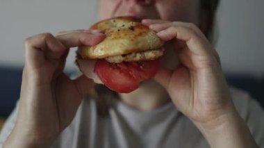 Fast food aşığı. Evdeki mutfakta etli ve domatesli hamburger yiyen mutlu kadın. Yüksek kaliteli FullHD görüntüler