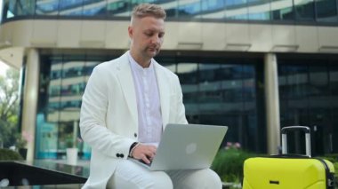 Odaklanmış genç bir işadamı bavulu olan bir yatırımcı iş merkezinin yanında oturuyor, bilgisayarında uzaktan kumanda ile çalışıyor. Yüksek kaliteli FullHD görüntüler