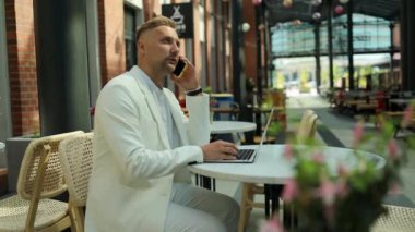 Mutlu Avrupalı işadamı akıllı telefondan bir telefon görüşmesi yapıyor, dışarıda şehir caddesindeki kafede oturup konuşuyor ve gülümsüyor. Yüksek kaliteli FullHD görüntüler