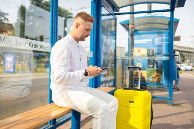 Kafkasyalı turist çantalı ve çantalı gezgin otobüs durağında cep telefonuyla bekliyor. 