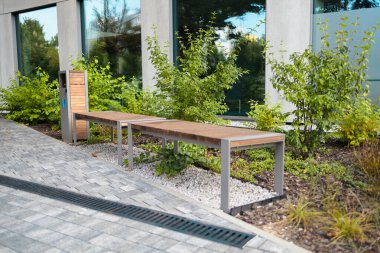 Yeşil çimenli asfalt yol, dekoratif çimenler ve modern ahşap bankların önünde kablodan yapılmış ve modern ofis binasının yakınlarındaki dinlenme alanında taşlarla doldurulmuş gabion ağaç küvetlerinin manzarası.