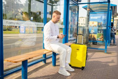 Kafkasyalı turist çantalı ve çantalı gezgin otobüs durağında cep telefonuyla bekliyor. 