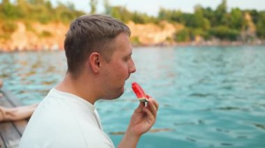 Şık giyinmiş yakışıklı genç bir adam bir dağ gölünün yanında oturur ve dinlenir. Adam iskelede lezzetli bir karpuz yiyor ve zevk alıyor..