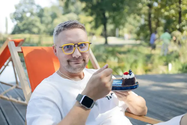 stock image Hungry Caucasian man in yellow glasses eating fresh sweet dessert enjoying taste at public cafe. Beautiful blonde male feeling pleasure with delicious food cupcake whipped cream at cozy coffeeshop. 