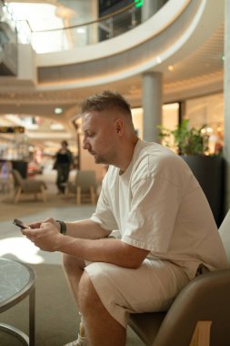  Yakışıklı, beyaz, şık giyinmiş, alışveriş merkezindeki yumuşak koltukta cep telefonu kullanan yetişkin bir adam. 