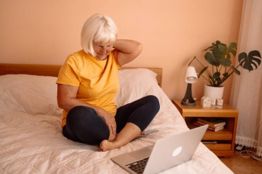 Yorgun, son sınıf öğrencisi, 60 'lı yaşlarda bir kadın sabah laptopuyla yatakta çalışıyor. Kadın yatakta oturuyor, sırtına ve boynuna dokunuyor, vücut ağrıları çekiyor, pijama giyiyor..