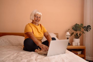 Üst düzey beyaz kadın dizüstü bilgisayarla çalışıyor, apartmandaki bir yatakta online alışveriş yapıyor.