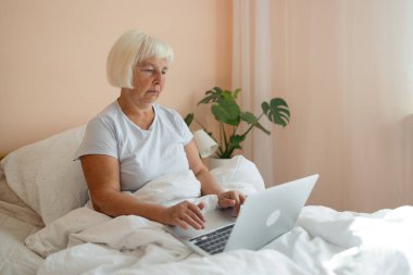 60 'lı yaşlarda evde yatağın üzerinde dizüstü bilgisayarı üzerinde çalışan mutlu ve güzel bir kadın. Serbest meslek, uzak çalışma, evde çalışma, online eğitim. Kişisel tecrit. 