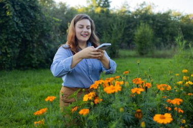 Bahçesinde çalışan mutlu bir beyaz kadın çiftçiyi telefon kullanarak ve fotoğraf çekerken çekmiş. Bahçıvan cep telefonu kullanıyor. Bahçe işleri, çiçek ve bitkilerle ilgilenmek ve büyümek..