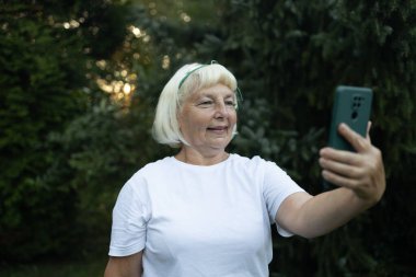 Beyaz tişörtlü, kameraya bakıp gülümseyen, akıllı telefondan selfie çeken mutlu yaşlı kadın portresi. Bulanık arkaplanda cadde