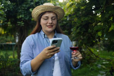 Hasır şapkalı ve telefonlu beyaz kadının portre görüntüsü üzüm bağında kırmızı şarap tadıyor. Başarılı üzüm hasadından sonra şarap içen mutlu şarap üreticisi, doğal şarap endüstrisi. Hasat zamanı