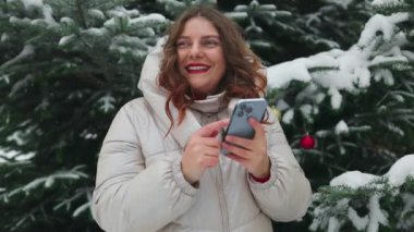 Karlı bir kış parkında cep telefonunu kullanan genç bir kadın. Soğuk kış mevsiminde, haberlere bakan ve dışarıda cep telefonuyla mesajlaşan kadınların yakınlaşması. İnsanlar araç gereç konsepti. Yüksek kalite FullHD