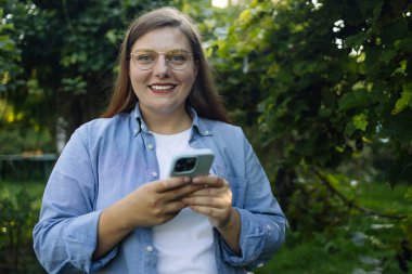  Beyaz kadın arka bahçede akıllı telefon kullanarak gülümsüyor. Genç bir kadın internette geziniyor, dışarıda mesajlaşıyor ve iletişim kuruyor. Seyahat, moda, blog konsepti. Yüksek kalite fotoğraf