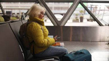 Havaalanındaki bankta oturan mutlu, güzel bayan gezgin, dijital akıllı telefon tutuyor, uçuşu bekliyor, arkadaşlarıyla telefon görüşmesi yapıyor, fotokopi odasına bakıyor. Yüksek kalite fotoğraf