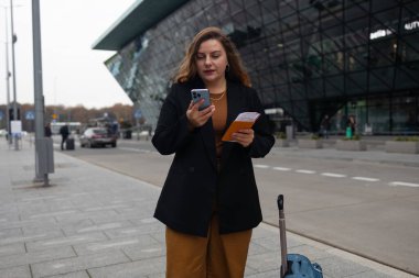 Havaalanından taksiye binen, 30 'lu yaşlarda, akıllı telefonlu, günlük kıyafetleri olan, gülümseyen bir bayan turist. Şehir caddesinde bir bavul. Seyahat kavramı.