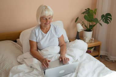 60 'lı yaşlarda evde yatağın üzerinde dizüstü bilgisayarı üzerinde çalışan mutlu ve güzel bir kadın. Serbest meslek, uzak çalışma, evde çalışma, online eğitim. Kişisel tecrit. 