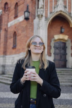 Genç ve şık bir kadın kahvesini mutlu bir ifadeyle içer. Şehir sokağında durur ve taksi bekler.
