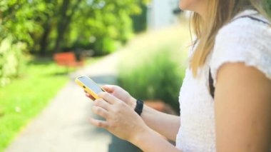 Yakın plan çekimde, açık havada bir bankta oturan ve akıllı telefonunu, insan yaşam tarzını ve kablosuz teknoloji kavramlarını kullanan bir kadın görülüyor. Yüksek kalite fotoğraf