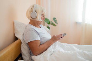 Kulaklık takan, akıllı telefon kullanan, teknolojiyi kucaklayan ve yatak odasında huzurlu bir anın tadını çıkaran yaşlı bir kadın.. 