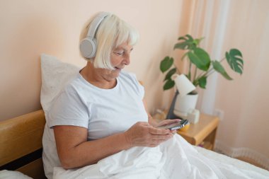 Kulaklık takan, akıllı telefon kullanan, teknolojiyi kucaklayan ve yatak odasında huzurlu bir anın tadını çıkaran yaşlı bir kadın.. 