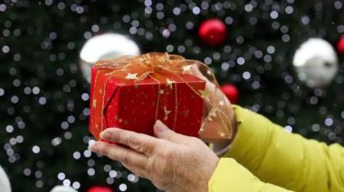 Kırmızı kurdeleli paketlenmiş yılbaşı hediyesi. Mutlu kadın ellerinde. Işıklı Noel ağacının arka planında. Mutlu noeller. Yüksek kaliteli FullHD görüntüler