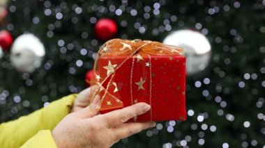 Kırmızı kurdeleli paketlenmiş yılbaşı hediyesi. Mutlu kadın ellerinde. Işıklı Noel ağacının arka planında. Mutlu noeller. Yüksek kaliteli FullHD görüntüler
