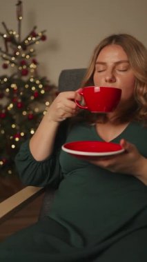 Kırmızı bardaktan aromalı kakao latte içen sevimli, sevimli, tatlı, neşeli bir kızın yakın plan portresi. Mutlu kadın evde rahatlıyor ve gülümsüyor. Yılbaşı Noel atmosfer dekorasyonuyla birlikte.