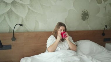 Beyaz pijamalı bir kadın güneşli sabah yatak odasında oturup kahve içiyor.. 