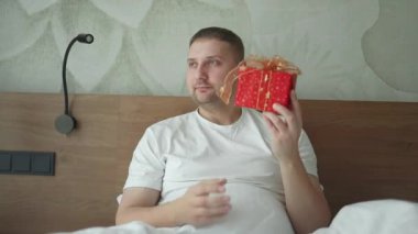 Yakışıklı beyaz bir adam sarılı hediyesini açıyor. Otelde bir yatakta mutlu ve heyecanlı hissediyor. Sevgililer Günü kutlamaları, yüksek kaliteli FullHD görüntüleri.