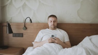 Happy Kafkas 'lı genç adam aramalar, sosyal ağlar, oteldeki bir yatakta online mobil uygulamalar için akıllı telefon kullanıyor. Yüksek kaliteli FullHD görüntüler