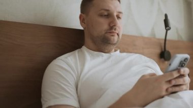 Happy Kafkas 'lı genç adam aramalar, sosyal ağlar, oteldeki bir yatakta online mobil uygulamalar için akıllı telefon kullanıyor. Yüksek kaliteli FullHD görüntüler