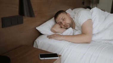 Telefondaki alarmı çalıştırdıktan sonra uyuyan adamı uyandırır. Yakışıklı, uykulu, beyaz bir adam yatakta uyuyor. Akıllı telefondan ne kadar zaman geçtiğine bakıyor. 