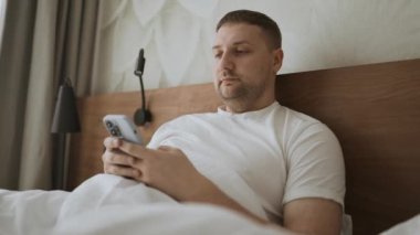 Happy Kafkas 'lı genç adam aramalar, sosyal ağlar, oteldeki bir yatakta online mobil uygulamalar için akıllı telefon kullanıyor. Yüksek kaliteli FullHD görüntüler