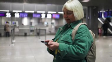Havaalanındaki kadın terminalde bekliyor. Uluslararası havaalanında yeşil ceketli, gülümseyen bir iş kadını, uçuşunu beklerken telefonunu kontrol ediyor.