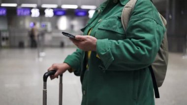 Havaalanındaki kadın terminalde bekliyor. Uluslararası havaalanında yeşil ceketli, gülümseyen bir iş kadını, uçuşunu beklerken telefonunu kontrol ediyor.