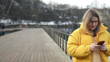 Kışlık sarı ceket giyen, akıllı telefonlu, göl kenarında yürüyen ve güzergahı kontrol eden Kafkasyalı genç bir turist kadın. Özgürlük, mutluluk, seyahat ve tatil konsepti, açık hava aktiviteleri