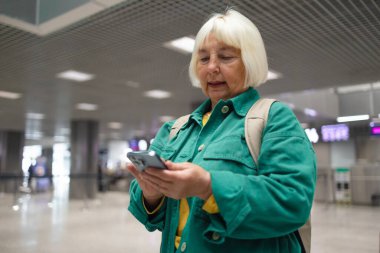 Havaalanındaki kadın terminalde bekliyor. Uluslararası havaalanında yeşil ceketli, gülümseyen bir iş kadını, uçuşunu beklerken telefonunu kontrol ediyor.
