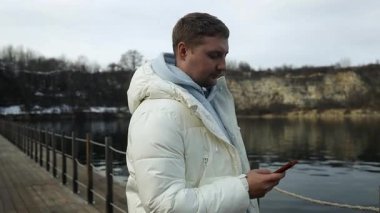  Soğuk kış günü, 30 'lu yaşlarda, nehrin kenarında telefonla dolaşan beyaz bir adam. Seyahat, sosyal medya veya kırsal bölgelerdeki maceralar hakkında bloglar için gölde insanlar, telefon veya bağlantı 