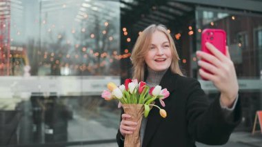 Şaşırmış genç bir kadın, taze çiçeklerle akıllı telefondan selfie çekerken son derece mutlu. Heyecanlı kız telefon ekranına bakıyor ve sevinçten havalara uçuyor. Yüksek kaliteli FullHD görüntüler