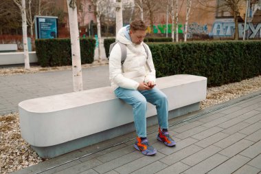 Şehir parkında bankta oturan, akıllı telefon kullanan, müzik dinleyen, akıllı telefonuna bakarken öksüren beyaz bir adam. Kış tatili Yüksek kalite fotoğraf