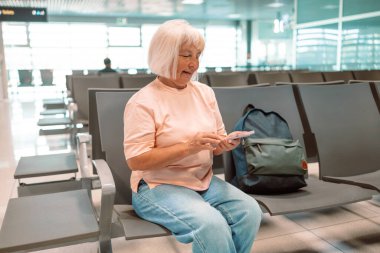 Üst düzey bir beyaz kadın turistin görüntüsü elinde akıllı telefonu ve pasaportuyla havaalanı terminalinde beklerken uçuş bilgilerini kontrol ediyor..