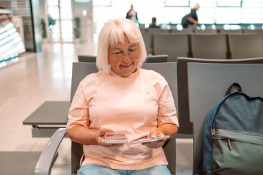 Üst düzey bir beyaz kadın turistin görüntüsü elinde akıllı telefonu ve pasaportuyla havaalanı terminalinde beklerken uçuş bilgilerini kontrol ediyor..