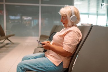 Havaalanı terminalinde tatil için uçağa binmeyi bekleyen 50 'li yaşlarda beyaz çekici bir kadın. Akıllı telefon kullanıyor, müzik dinlemek için kulaklık takıyor yanında bavul taşıyor.