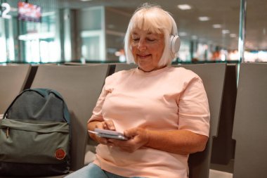 Havaalanı terminalinde tatil için uçağa binmeyi bekleyen 50 'li yaşlarda beyaz çekici bir kadın. Akıllı telefon kullanıyor, müzik dinlemek için kulaklık takıyor yanında bavul taşıyor.