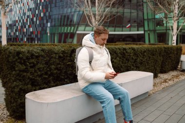 Şehir parkında bankta oturan, akıllı telefon kullanan, müzik dinleyen, akıllı telefonuna bakarken öksüren beyaz bir adam. Kış tatili Yüksek kalite fotoğraf