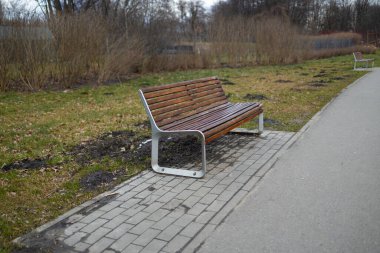Yürüyüş yolu boyunca ahşap bir bankın olduğu huzurlu bir park sahnesi. Kamu parkındaki ağaçların ve yeşilliğin tadını çıkarırken bank dinlenmek için bir yer sağlıyor.