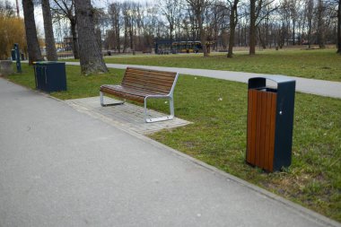 Ahşap bir bank, çöp bidonları ve yeşil park ortamının yer aldığı iyi korunmuş bir park sahnesi. Yol, arka planda ağaçlarla parkın içinden geçiyor. Huzurlu bir atmosfer sunuyor.