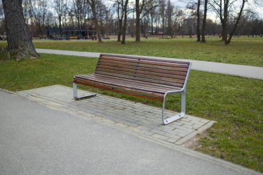Yürüyüş yolu boyunca ahşap bir bankın olduğu huzurlu bir park sahnesi. Kamu parkındaki ağaçların ve yeşilliğin tadını çıkarırken bank dinlenmek için bir yer sağlıyor.
